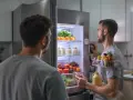 Deux hommes devant un frigo rempli de fruits et légumes. L'un tient une corbeille de fruits, l'autre un téléphone. Mon frigo ne refroidit plus mais congélateur fonctionne.