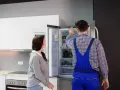 Un technicien en salopette bleue vérifie un frigo qui claque, tandis qu'une femme observe attentivement.