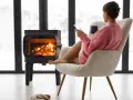 Femme assise près d'un poêle à bois, tenant une tasse et un téléphone.