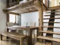 Une salle à manger rustique avec une table en bois, des chaises et un banc, sous une mezzanine maison ancienne aux poutres apparentes. L'escalier en bois brut complète le décor.