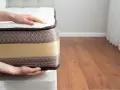 Mains posées sur un matelas, montrant sa bonne épaisseur. Le tissu matelassé marron et la bande beige suggèrent un confort optimal.