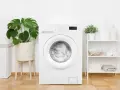 Machine à laver blanche, prête pour un cycle. La profondeur de son tambour est idéale pour une famille. À côté, un panier à linge et des plantes vertes.