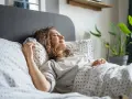 Une femme souriante se repose dans un lit douillet. Elle semble heureuse, une belle illustration de comment choisir une bonne literie pour un sommeil réparateur.
