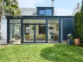 Une extension de maison moderne avec une immense baie vitrée s'ouvrant sur un jardin verdoyant. Une palme se dresse à gauche.
