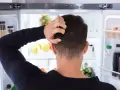Un homme se gratte la tête devant un réfrigérateur ouvert, plein de nourriture. Il semble réfléchir à un réglage frigo.