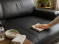 Nettoyer simili cuir bicarbonate : une main essuie un canapé noir avec un chiffon. Un spray et un bol de mousse sont à proximité.