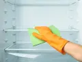 Main d'une personne portant un gant orange, essuyant une étagère de réfrigérateur vide avec une éponge verte. Le nettoyage du frigo est terminé.