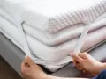 Mains ajustant les élastiques d'un surmatelas blanc et moelleux. Un surmatelas améliore le confort et la protection de votre matelas.