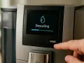 Une main appuie sur le bouton "Start" d'une machine Nespresso. L'écran affiche "Descaling" avec une barre de progression, indiquant le début du détartrage.