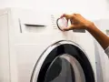 Main d'une personne ajustant le sélecteur d'un sèche-linge, choisissant un programme comme "Coton" ou "Laine". Les dimensions de la machine à laver sont standard.
