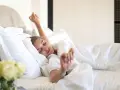 Une femme se réveille, s'étirant dans un lit douillet. La bonne literie blanche et les oreillers moelleux invitent à la détente.