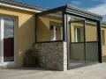 Une véranda moderne avec murs en pierre et grandes baies vitrées permet une entrée directe dans le séjour.