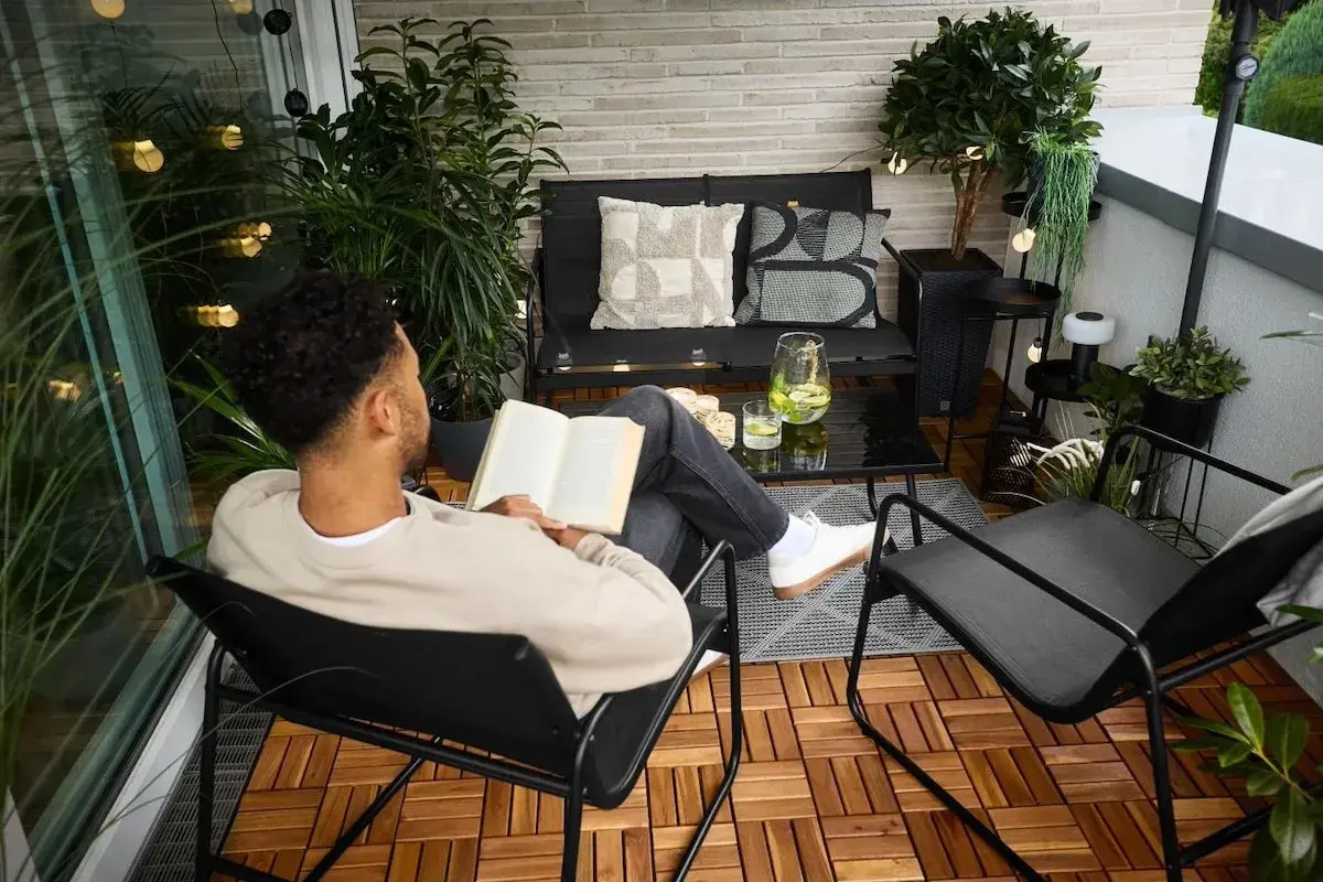 Un homme lit un livre sur un balcon aménagé, profitant d'un moment de détente. La deco balcon comprend des plantes, des coussins et des guirlandes lumineuses.