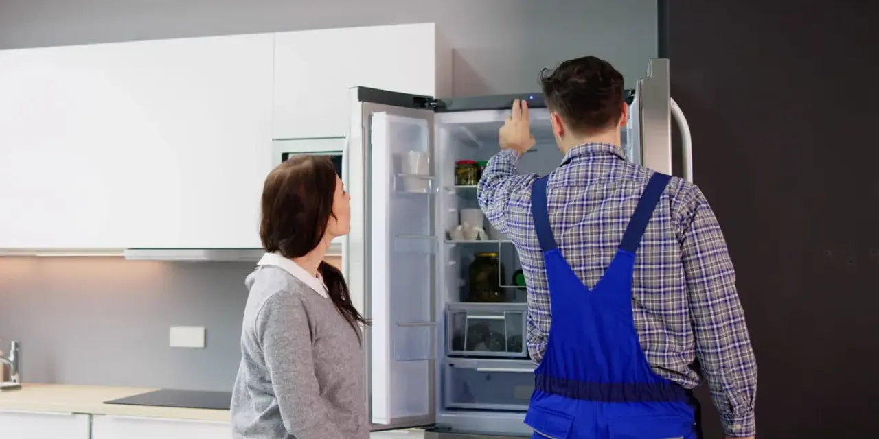 Un technicien en salopette bleue vérifie un frigo qui claque, tandis qu'une femme observe attentivement.