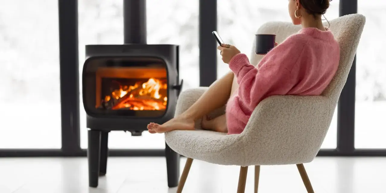 Femme assise près d'un poêle à bois, tenant une tasse et un téléphone.
