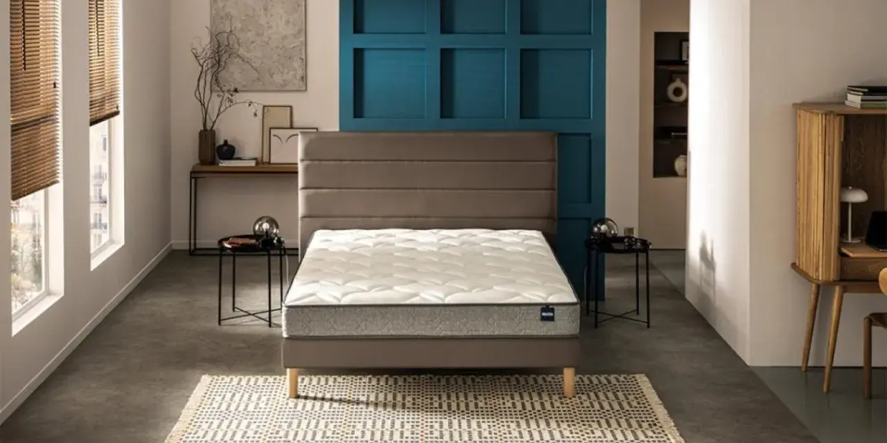Un lit moderne avec un matelas blanc et un sommier gris, sur un tapis géométrique. Le prix moyen d'un bon matelas est un investissement pour le confort.