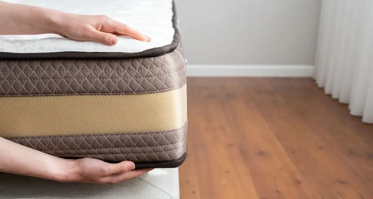 Mains posées sur un matelas, montrant sa bonne épaisseur. Le tissu matelassé marron et la bande beige suggèrent un confort optimal.
