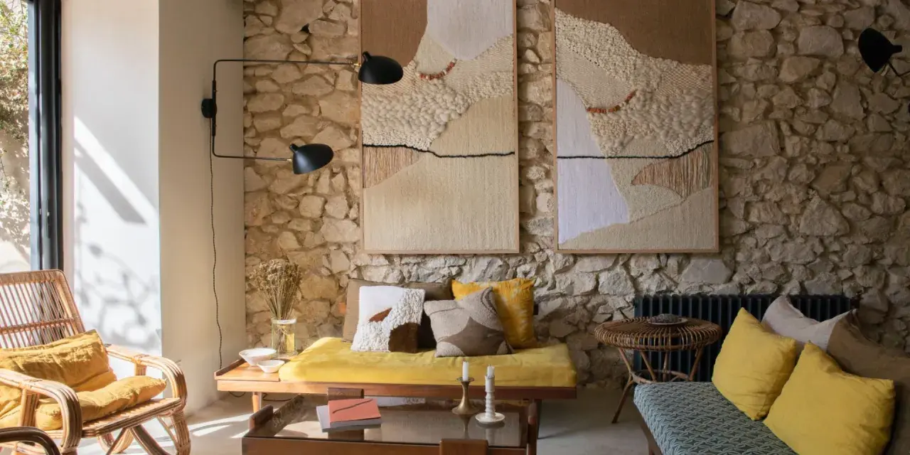 Salon avec mur en pierre, deux tapisseries texturées, fauteuil en rotin, banquette jaune et coussins aux motifs ethniques. Une ambiance deco campagne boheme chaleureuse.