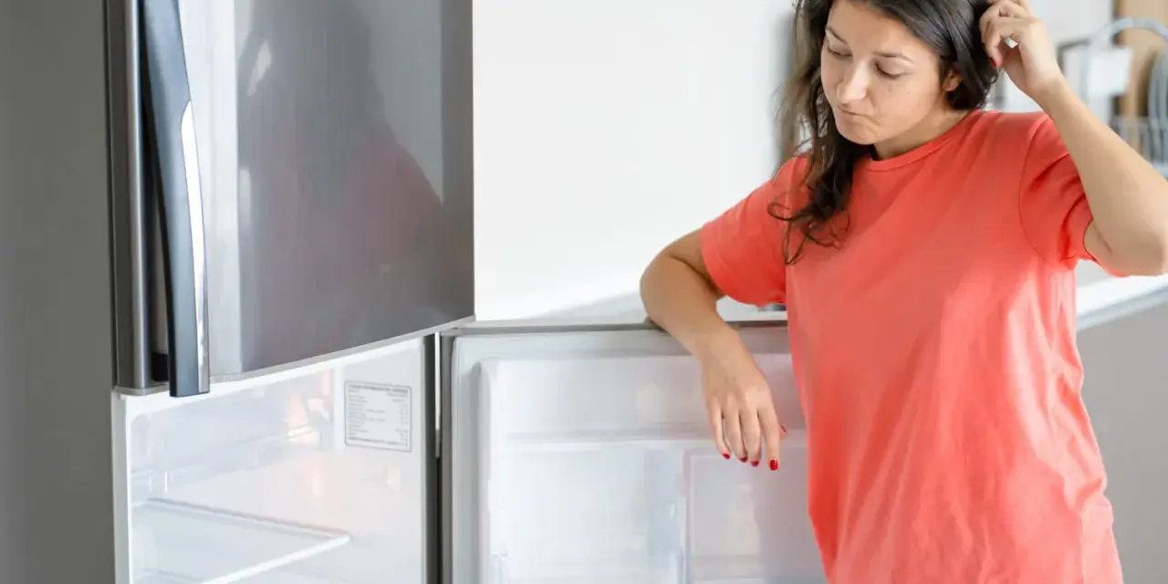 Une femme perplexe regarde son frigo vide, qui fait un bruit étrange. Elle se gratte la tête, se demandant ce qu'elle va manger.