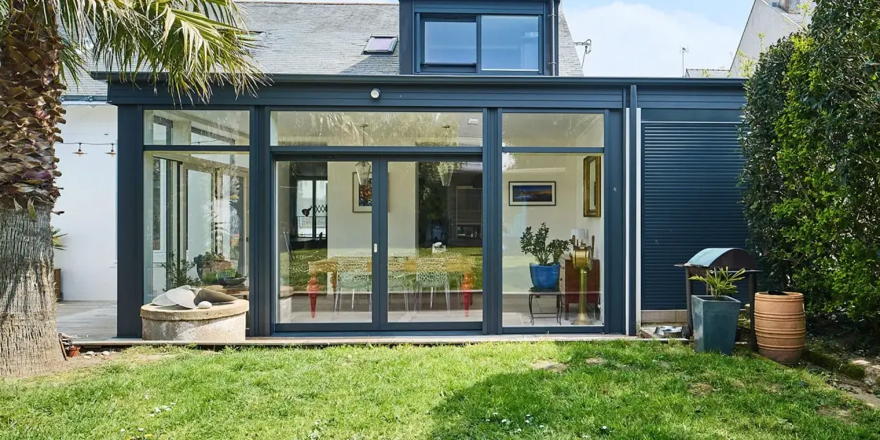 Une extension de maison moderne avec une immense baie vitrée s'ouvrant sur un jardin verdoyant. Une palme se dresse à gauche.
