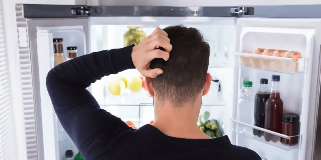 Un homme se gratte la tête devant un réfrigérateur ouvert, plein de nourriture. Il semble réfléchir à un réglage frigo.