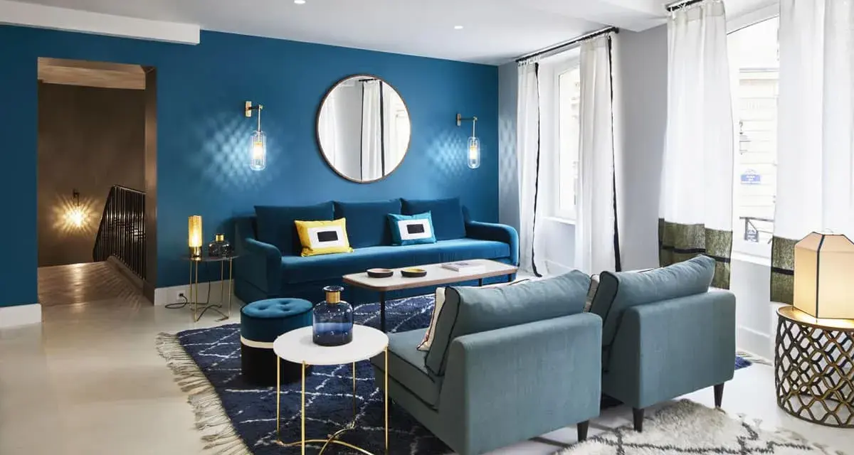Salon moderne avec mur bleu canard, canapé velours, fauteuils et tapis assortis. Le jaune vif des coussins dynamise l'ensemble.