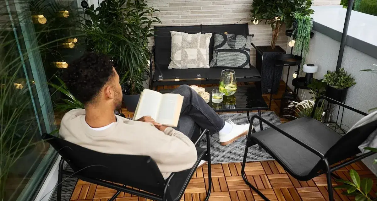 Un homme lit un livre sur un balcon aménagé, profitant d'un moment de détente. La deco balcon comprend des plantes, des coussins et des guirlandes lumineuses.