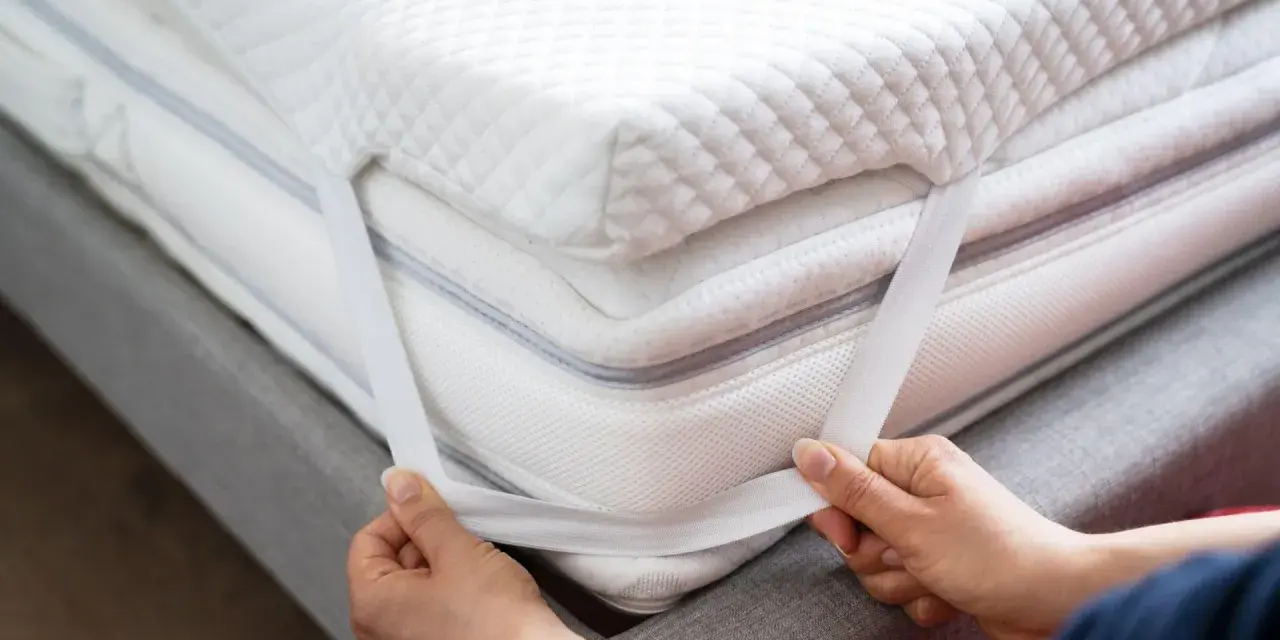 Mains ajustant les élastiques d'un surmatelas blanc et moelleux. Un surmatelas améliore le confort et la protection de votre matelas.