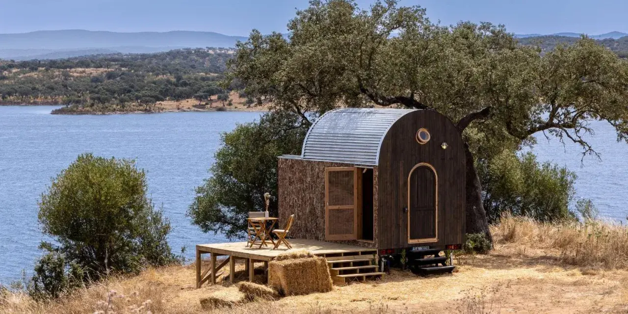 Une caravane transformée en cabane insolite, avec une terrasse en bois, au bord d'un lac paisible.