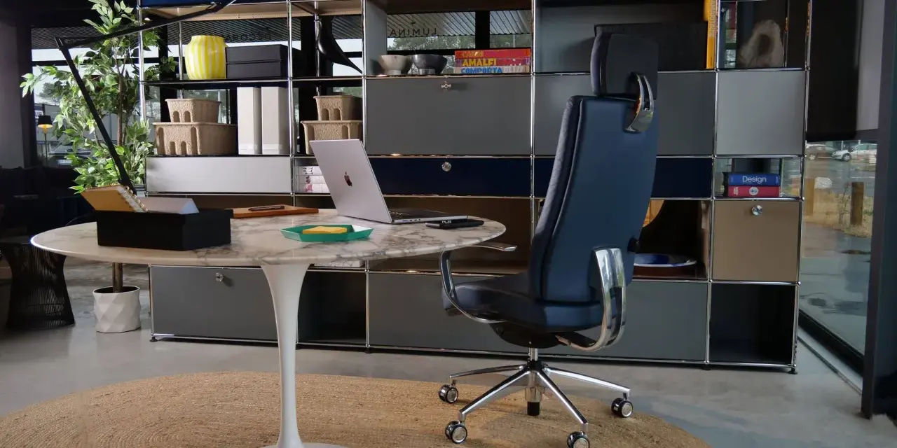 Un fauteuil de bureau design bleu marine, un bureau rond en marbre et un ordinateur portable créent un espace de travail élégant.