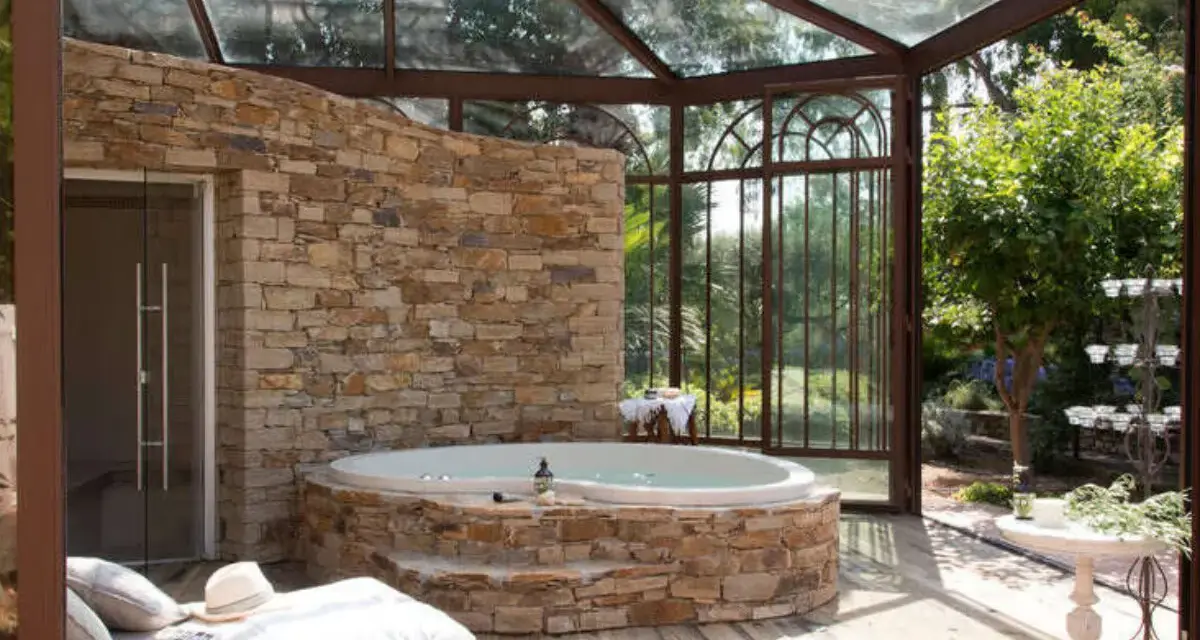 Un spa dans une cave, avec un jacuzzi en pierre, une banquette et une verrière donnant sur un jardin luxuriant.