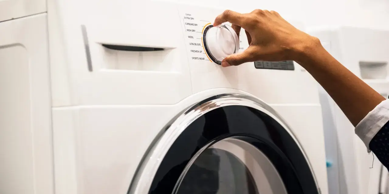Main d'une personne ajustant le sélecteur d'un sèche-linge, choisissant un programme comme "Coton" ou "Laine". Les dimensions de la machine à laver sont standard.