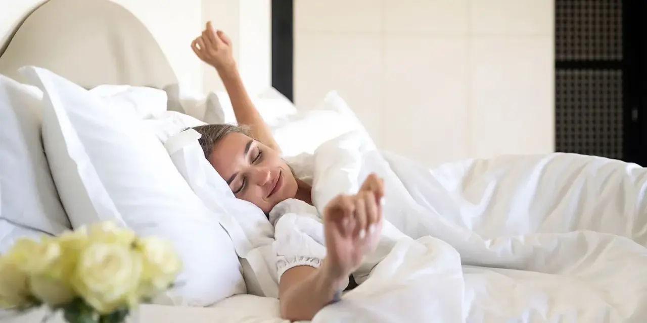 Une femme se réveille, s'étirant dans un lit douillet. La bonne literie blanche et les oreillers moelleux invitent à la détente.