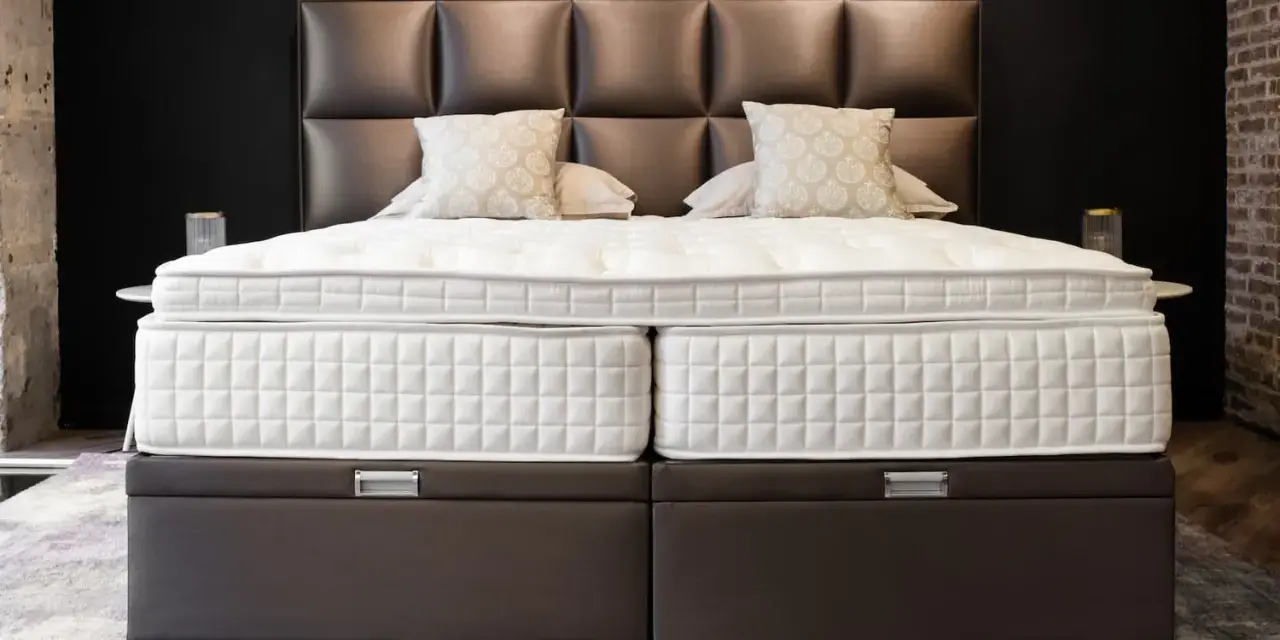 Lit, deux matelas blancs sur des sommiers marron. Le matelas trop grand pour sommier crée un léger débord.