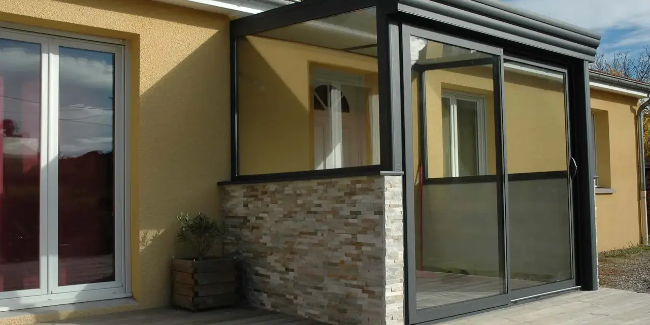 Une véranda moderne avec murs en pierre et grandes baies vitrées permet une entrée directe dans le séjour.