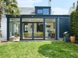 Une extension de maison moderne avec une immense baie vitrée s'ouvrant sur un jardin verdoyant. Une palme se dresse à gauche.