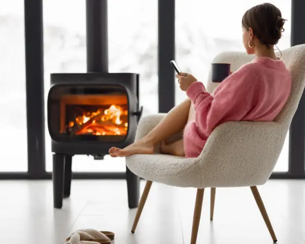 Femme assise près d'un poêle à bois, tenant une tasse et un téléphone.
