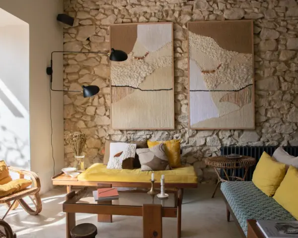 Salon avec mur en pierre, deux tapisseries texturées, fauteuil en rotin, banquette jaune et coussins aux motifs ethniques. Une ambiance deco campagne boheme chaleureuse.