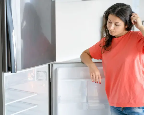 Une femme perplexe regarde son frigo vide, qui fait un bruit étrange. Elle se gratte la tête, se demandant ce qu'elle va manger.
