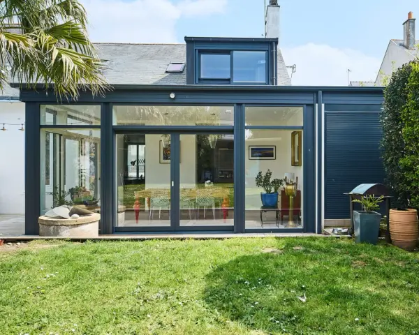 Une extension de maison moderne avec une immense baie vitrée s'ouvrant sur un jardin verdoyant. Une palme se dresse à gauche.