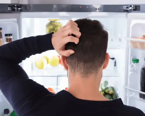 Un homme se gratte la tête devant un réfrigérateur ouvert, plein de nourriture. Il semble réfléchir à un réglage frigo.