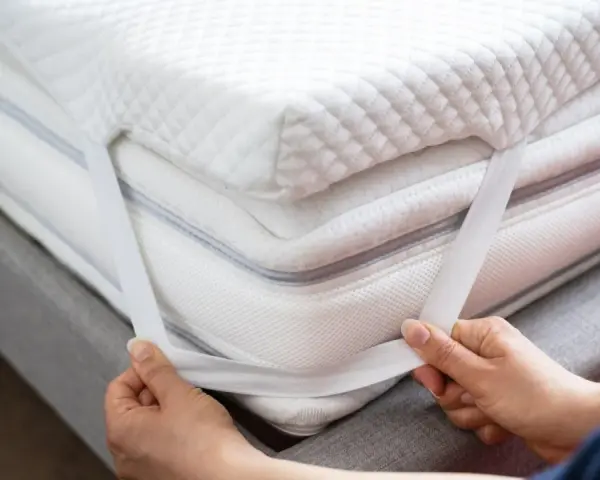 Mains ajustant les élastiques d'un surmatelas blanc et moelleux. Un surmatelas améliore le confort et la protection de votre matelas.