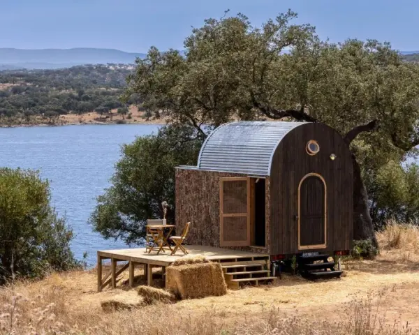 Une caravane transformée en cabane insolite, avec une terrasse en bois, au bord d'un lac paisible.