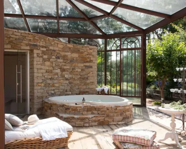 Un spa dans une cave, avec un jacuzzi en pierre, une banquette et une verrière donnant sur un jardin luxuriant.