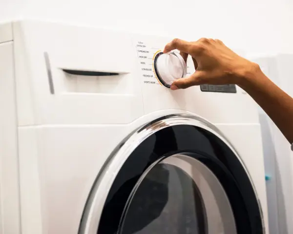 Main d'une personne ajustant le sélecteur d'un sèche-linge, choisissant un programme comme "Coton" ou "Laine". Les dimensions de la machine à laver sont standard.