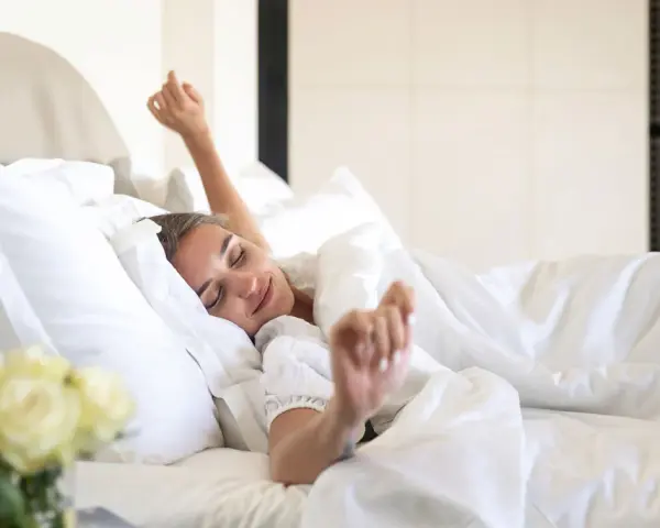Une femme se réveille, s'étirant dans un lit douillet. La bonne literie blanche et les oreillers moelleux invitent à la détente.