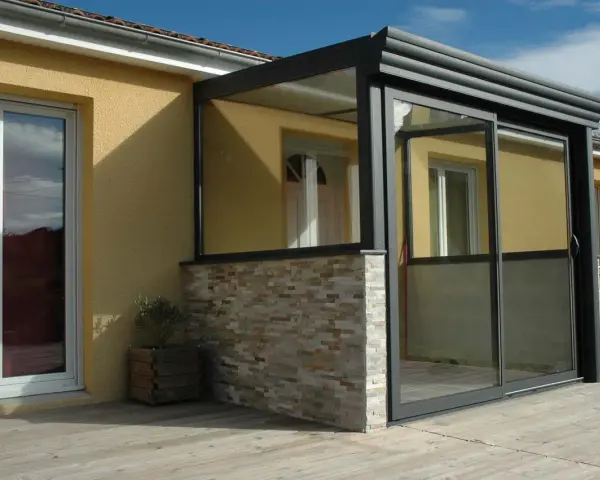 Une véranda moderne avec murs en pierre et grandes baies vitrées permet une entrée directe dans le séjour.
