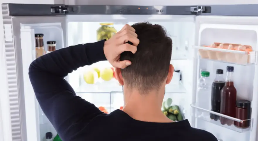Un homme se gratte la tête devant un réfrigérateur ouvert, plein de nourriture. Il semble réfléchir à un réglage frigo.