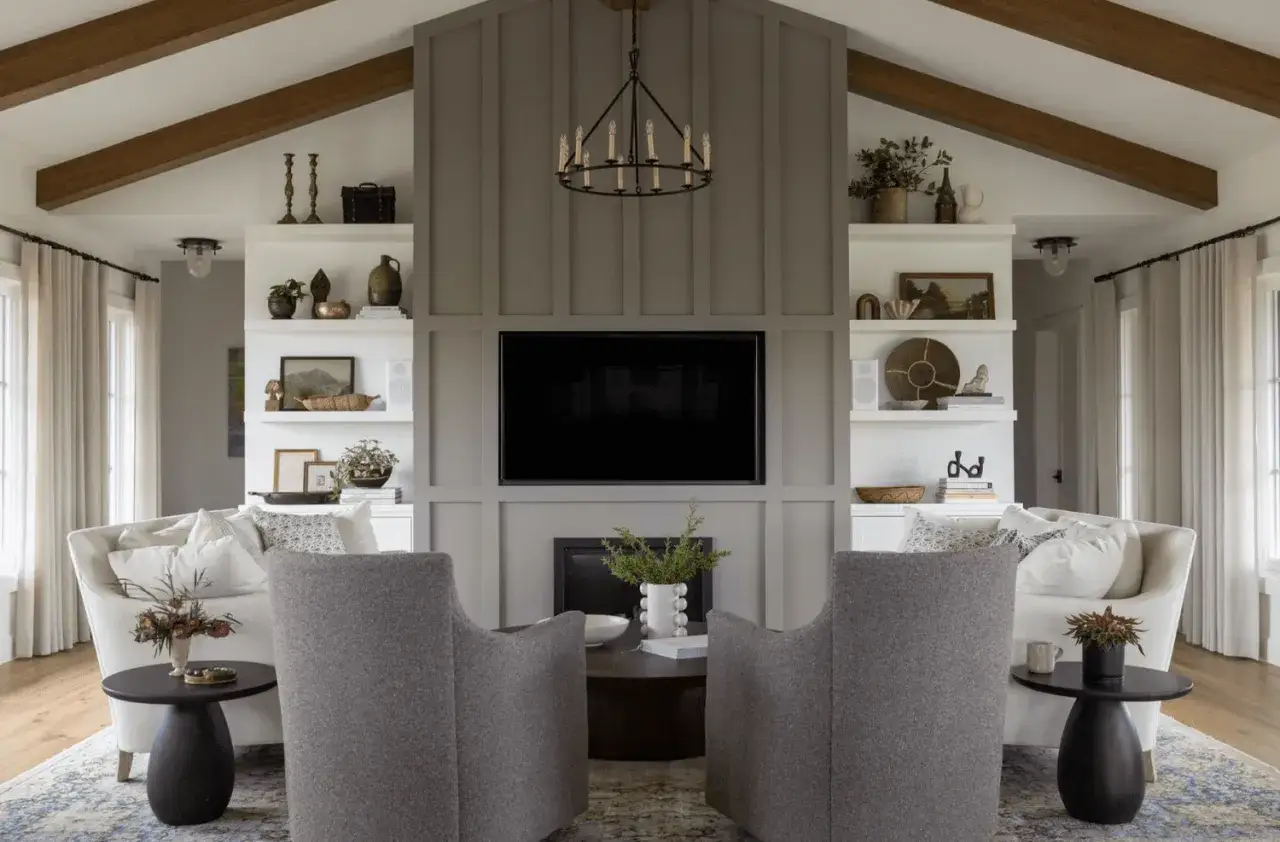 Salon moderne avec poutre apparente. Deux fauteuils gris font face à une cheminée et une télévision, encadrés par des étagères.