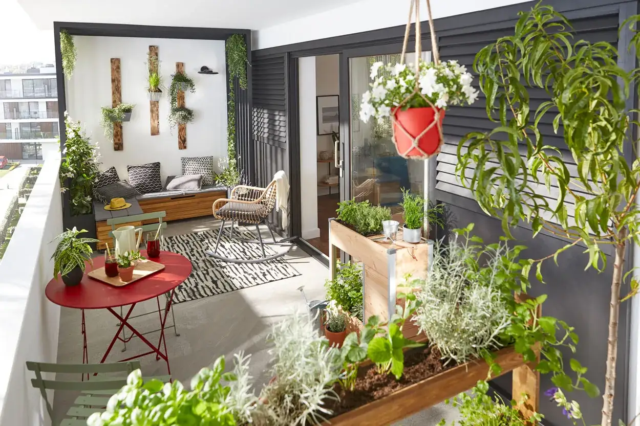 Un balcon aménagé avec une banquette, un fauteuil, une table rouge et de nombreuses plantes. Une belle deco balcon pour profiter du soleil.